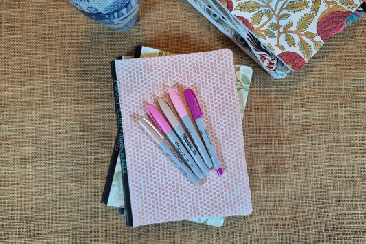 Stack of handwritten journals with pink and purple pens on a burlap surface