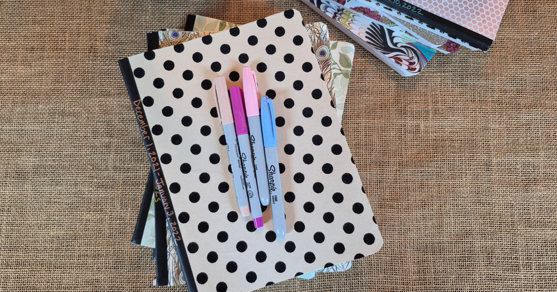 A polka-dot journal and colorful markers rest on a stack of notebooks, ready for a daily writing practice.