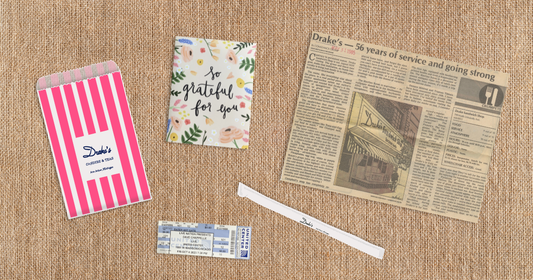 A flat lay of keepsakes on a burlap surface, including a pink striped bag, a gratitude card, a vintage newspaper clipping, and a worn straw — small things kept for the feelings they hold.